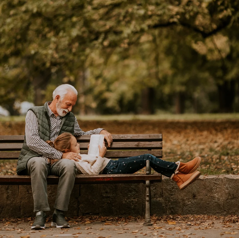 Ein älterer Mann und ein junges Mädchen lesen gemeinsam auf einer Parkbank im herbstlichen Park.