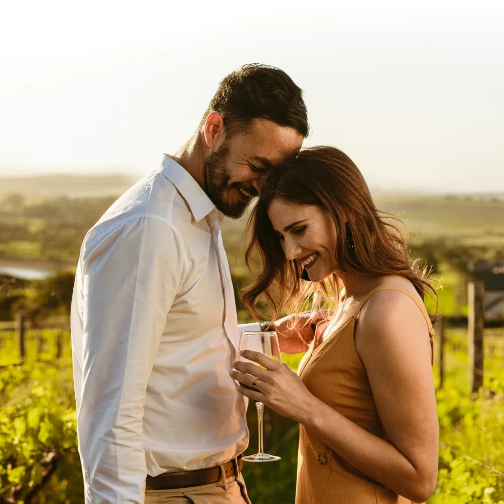 Ein lächelndes Paar steht mit einem Glas Wein in einem sonnigen Weinberg.